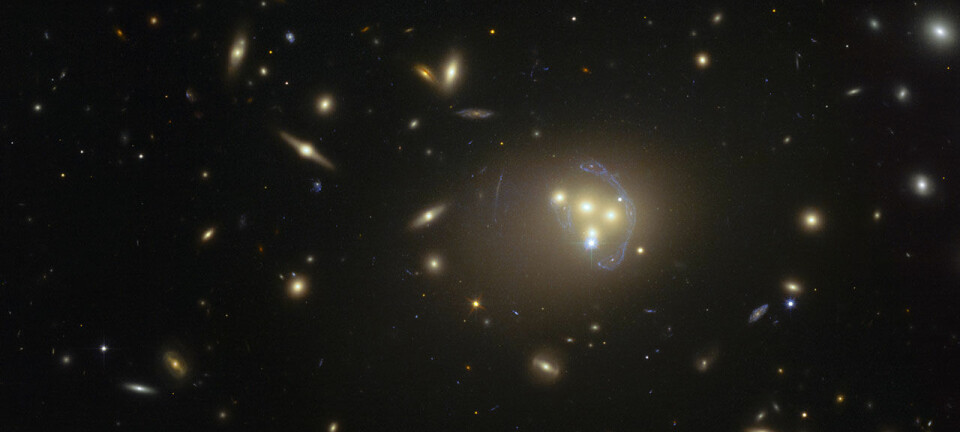 Galaksehopen Abell 3827 ligger omtrent midt i bildet. Legg merke til at lyset fra en fjern galakse må gjennom denne galaksekollisjonen, og selve lyset blir bøyd og strukket på grunn av de kraftige gravitasjonsfeltene. Du ser noe av lyset som en avlang banan til høyre. (Foto: ESO)