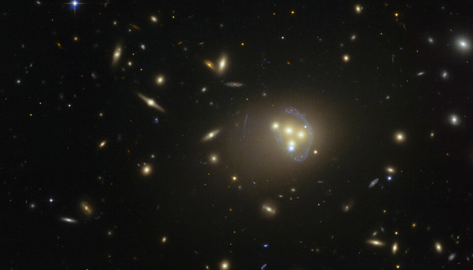 Galaksehopen Abell 3827 ligger omtrent midt i bildet. Legg merke til at lyset fra en fjern galakse må gjennom denne galaksekollisjonen, og selve lyset blir bøyd og strukket på grunn av de kraftige gravitasjonsfeltene. Du ser noe av lyset som en avlang banan til høyre. (Foto: ESO)