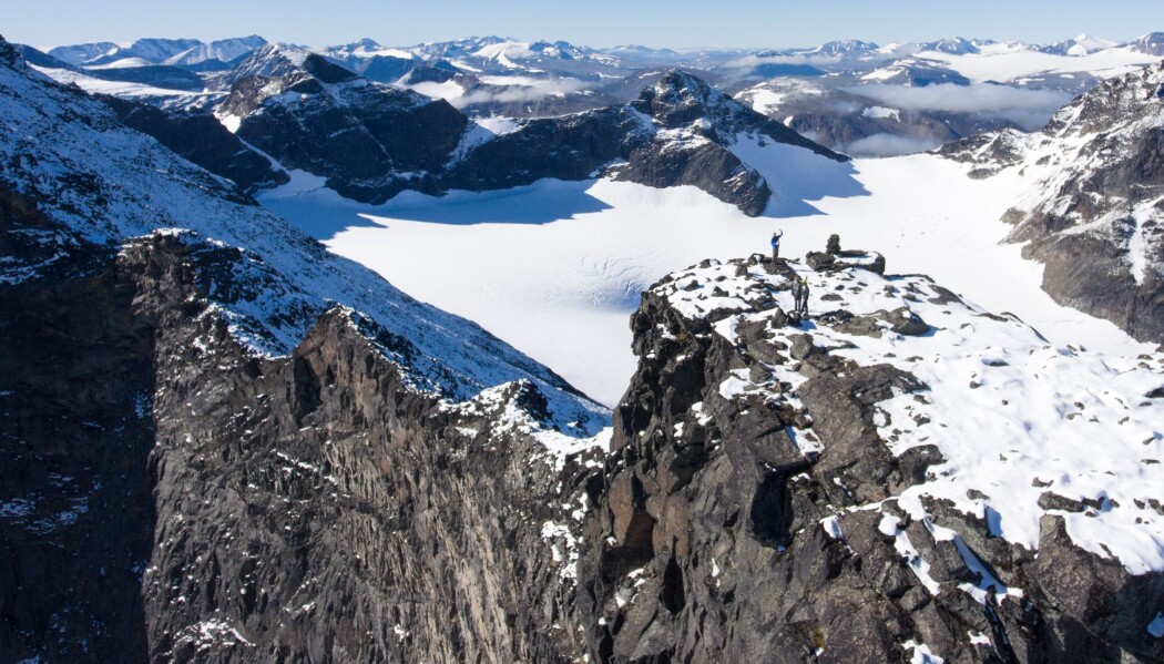 Fra en av toppene av Ähpar-massivet vinker Hans Jørgen Kjøll til helikopteret. Også her ligger mørke striper med lavastein på tvers i fjellveggen. Fra en av toppene av Ähpar-massivet vinker Hans Jørgen Kjøll til helikopteret. Også her ligger mørke striper med lavastein på tvers i fjellveggen.