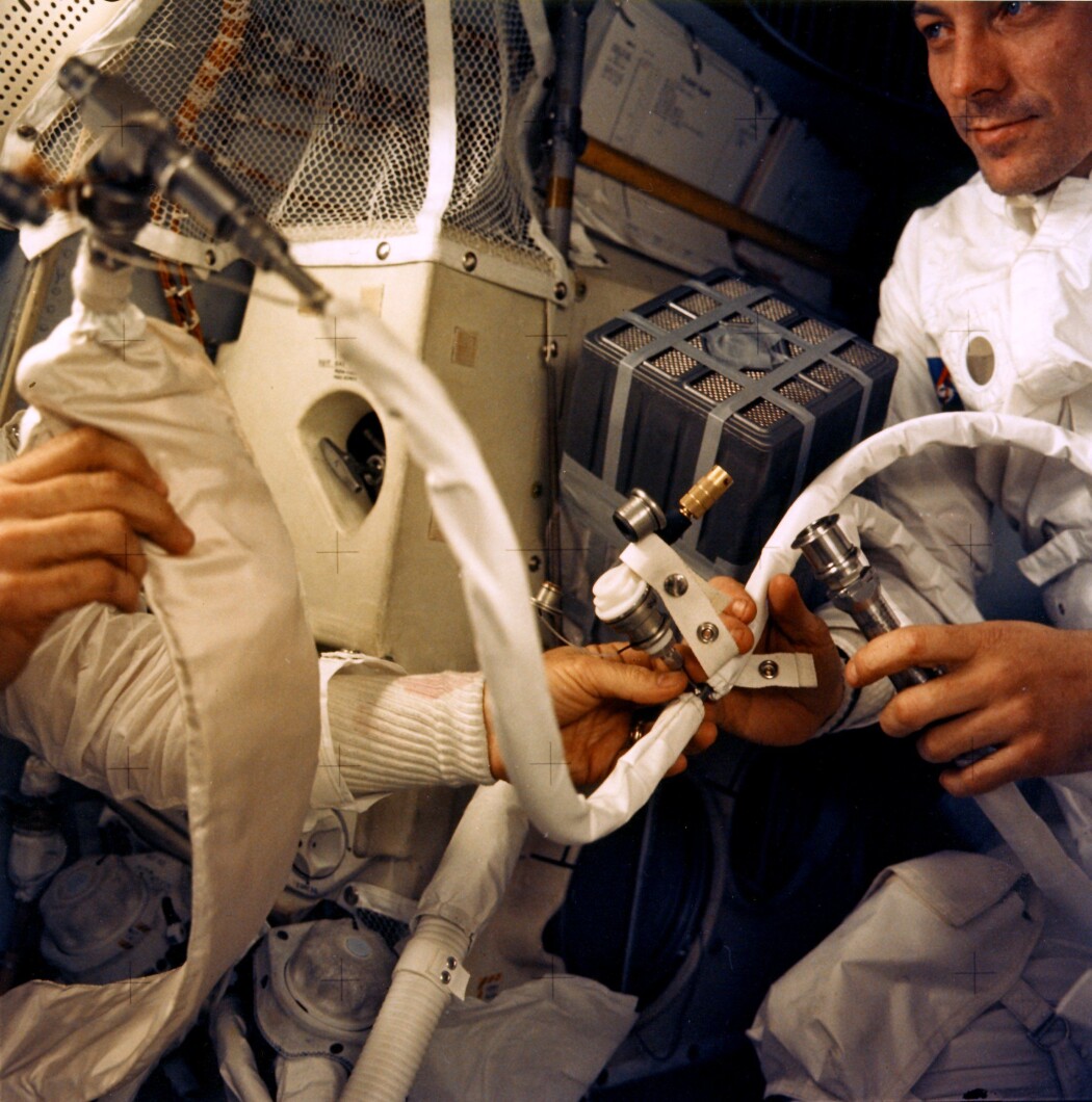 Astronautene ombord på Apollo 13 under ferden. Til høyre ser du John Swigert, og i midten av bildet er den berømte, improviserte filterboksen som skulle rense lufta i modulen for CO2. Astronautene ombord på Apollo 13 under ferden. Til høyre ser du John Swigert, og i midten av bildet er den berømte, improviserte filterboksen som skulle rense lufta i modulen for CO2.