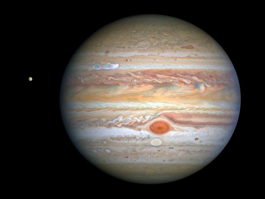 Jupiter som vi kjenner den, med den karakteristiske røde flekken, en storm som er 2–3 ganger større enn jorden, samt en ny, hvit stormsky som er synlig i den øvre siden av planeten. Til venstre for Jupiter ser vi ismånen Europa.