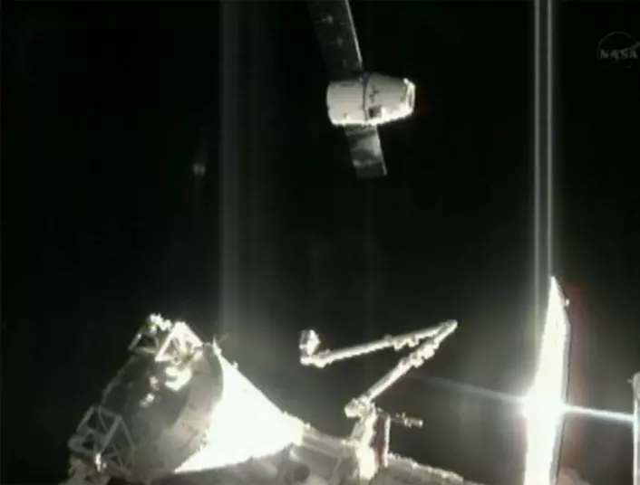 Romkapselen Dragon ankommer Den internasjonale romstasjonen 10.oktober 2012 med den første ordinære lasten. Nederst sees robotarmen Canadarm2, som skal strekkes ut for å fange inn Dragon. (Foto: NASA)