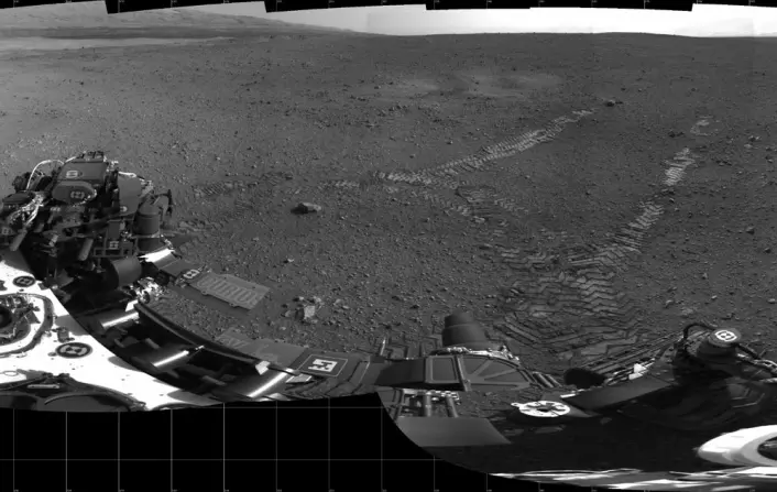 En mosaikk av småbilder tatt 22. august viser de første hjulsporene til kjøretøyet Curiosity på Mars. En smal stein, 89 centimeter i diameter, kan sees der det venstre sporet begynner. Ingeniørene mener at den lå under bakhjulet før kjøringen startet. Blåsemerker fra rakettmotorene som ble brukt under landingen kan sees på begge sider av sporene. (Foto: NASA/JPL)
