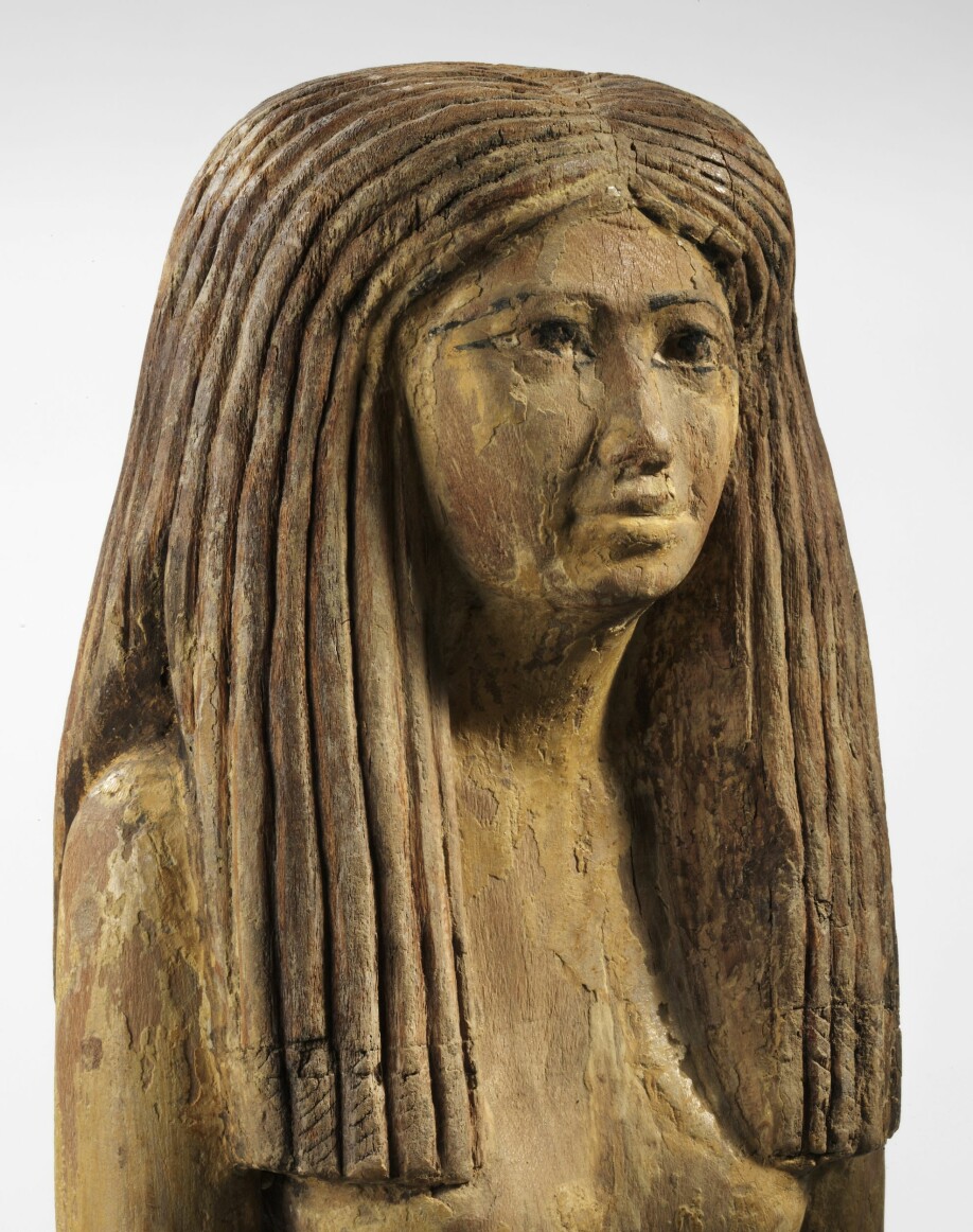 Trestatue av egyptisk kvinne, 1900–1800 f.Kr. Hun har en stor parykk, satt som brede lokker. Ved midtskillen ser vi en liten strek som viser et tørkle under parykken.