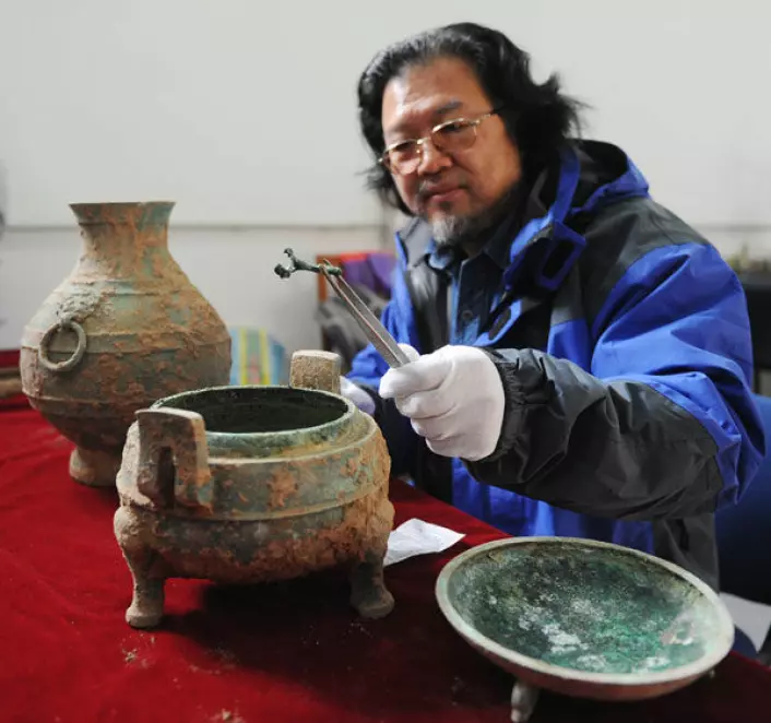 Arkeolog Liu Daiyun med gryte funnet i en 2400 år gammel grav ved Kinas gamle hovedstad Xian. Gryta inneholder dyrebein med irrgrønn patina. På bildet sees også en bronsekrukke som inneholdt en væske som kan være vin. (Foto: Zhai Xiaoxue/Xinhua/Photoshot)