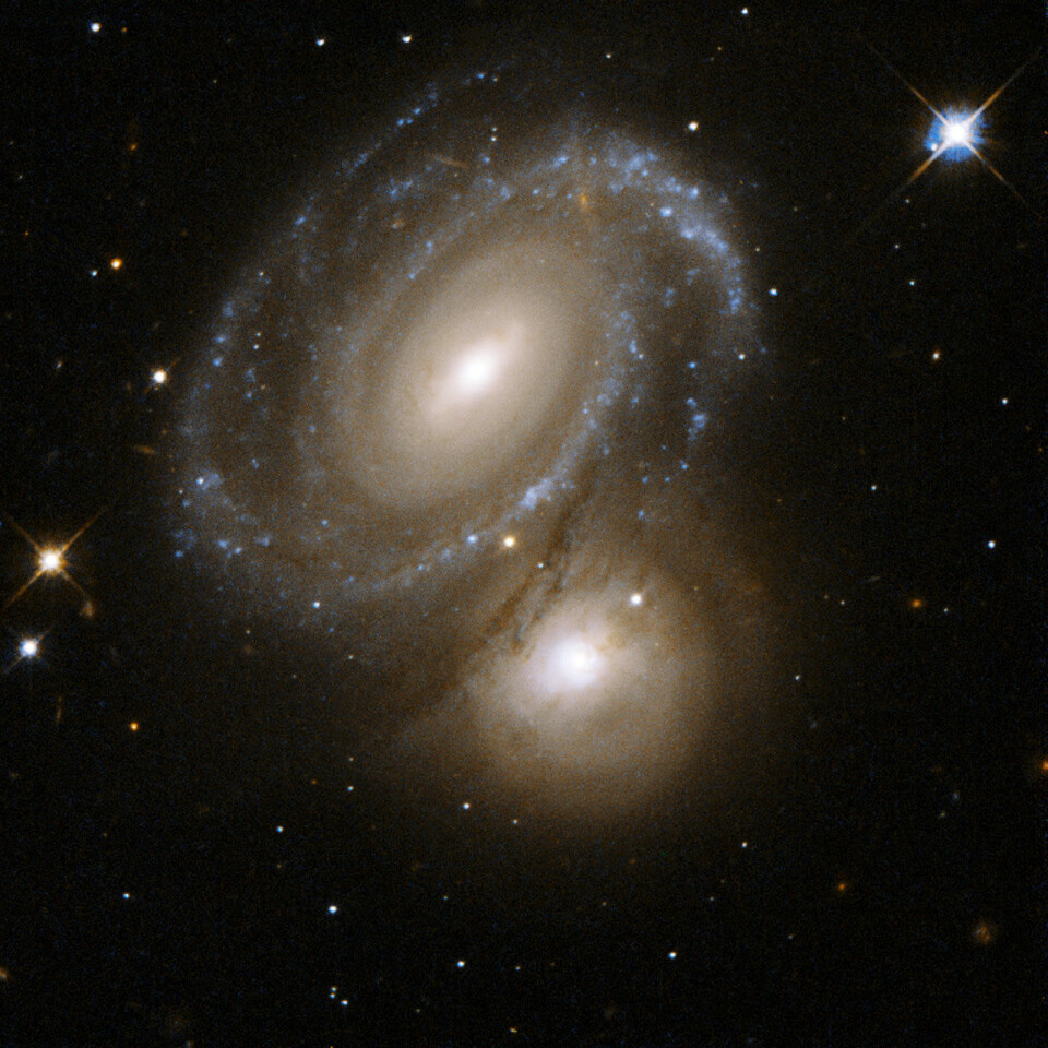 'Den nesten perfekt symmetriske galaksen i forgrunnen dekker, og er delvis opplyst av, den bakenforliggende galaksen. Sistnevnte galakse var tidligere klassifisert som en elliptisk galakse. Men de nye bildene fra Hubbleteleskopet avslører at det dreier seg om en spiralgalakse, med stjerner i knuteformasjoner. Galaksene befinner seg i stjernebildet Dorado, omlag 350 millioner lysår unna. (Foto: NASA/ESA)'