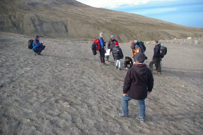 Turistene flokker seg rundt ekspedisjonsleder Jørn Hurum mens han gestikulerer og forteller om feltlivet og hvordan det er å drive en utgraving. (Foto: Bjørnar Kjensli)