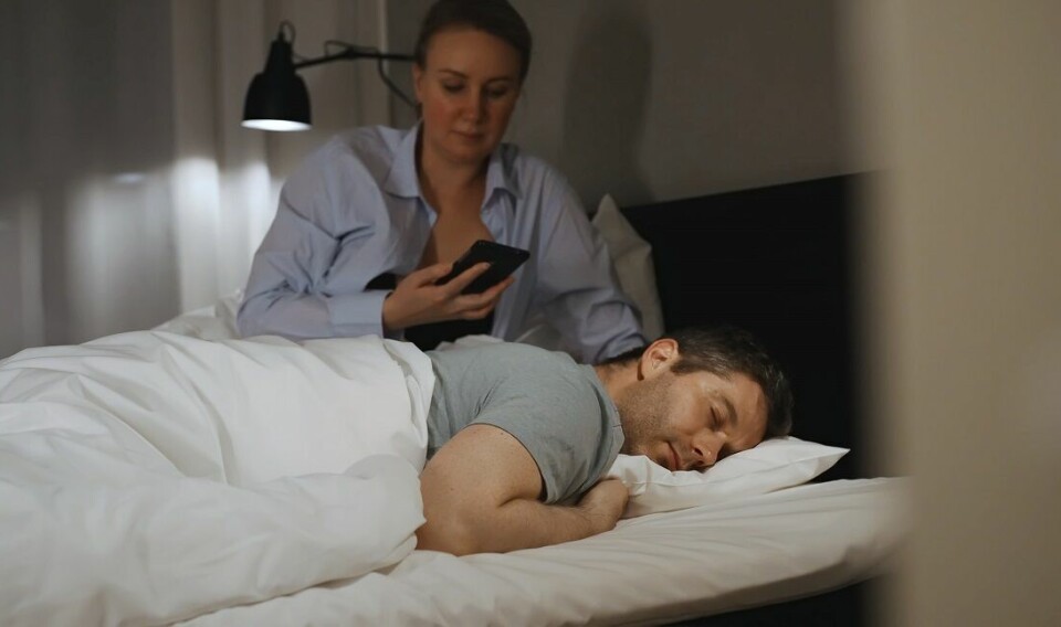 Kvinne sjekker telefon mens mann sover ved siden av.