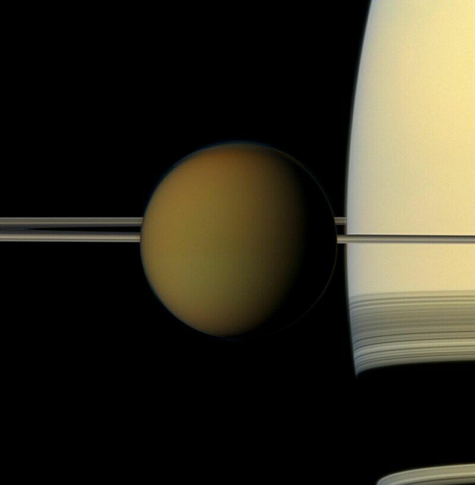 Titan sett i ekte farger av Cassini-sonden. I bakgrunnen ser du Saturn og ringen sett nesten rett på siden.