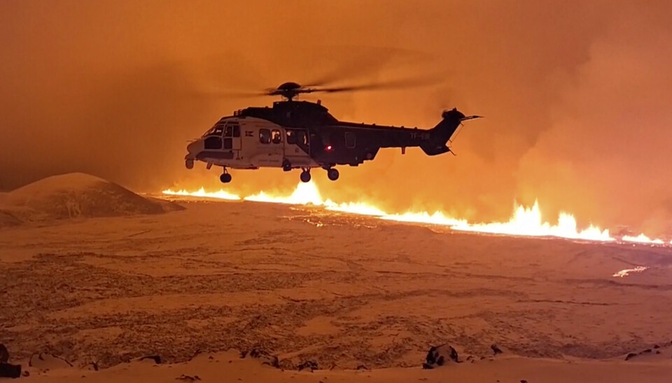 Helikopter svever over landskapet, en lang stripe av flammer strekker seg opp mot himmelen, der lavaen kommer ut av bakken.