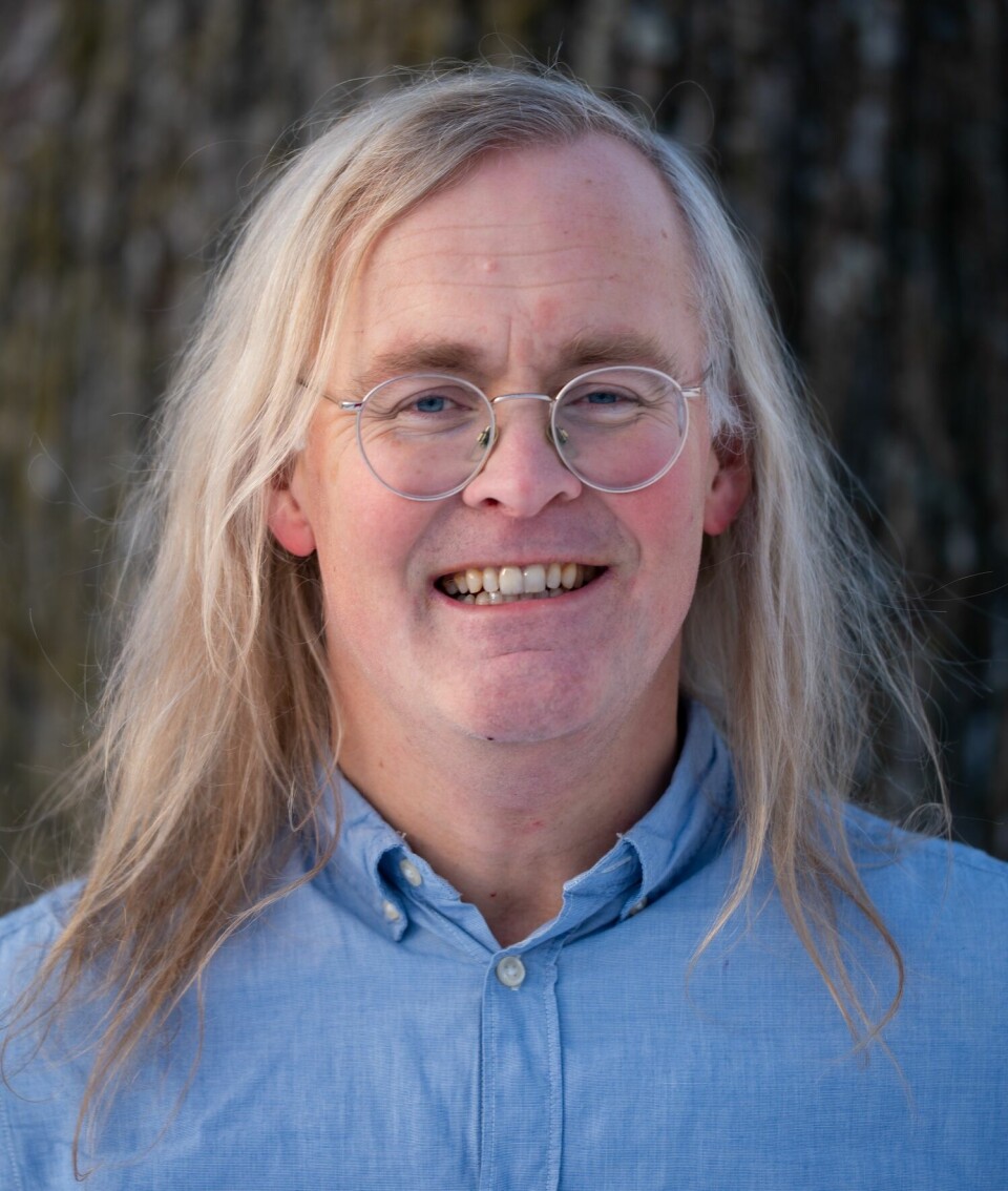 Portrait of a man with long blonde hair, round glasses, in a blue shirt.