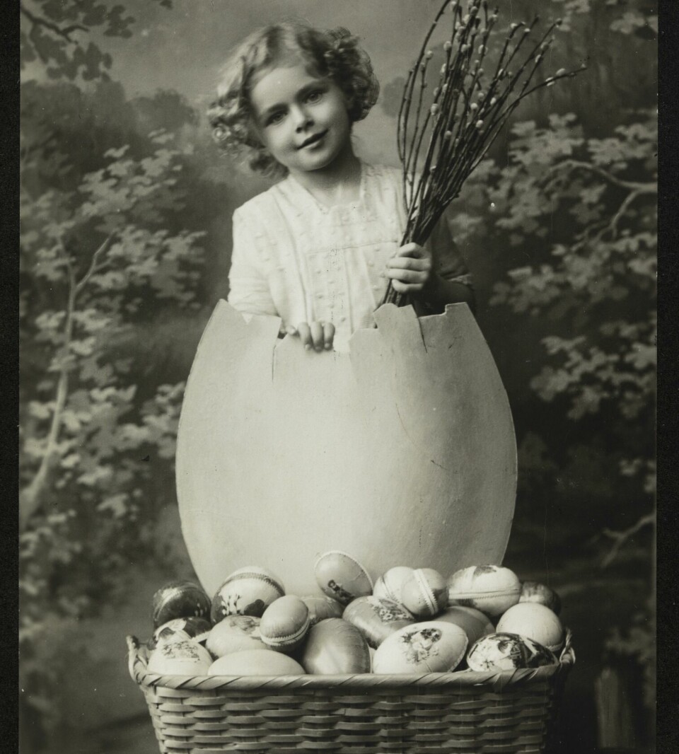 Et postkort i svart/hvitt som viser en liten jente som kommer ut av et stort egg. Ved føttene hennes står en kurv full av påskeegg.