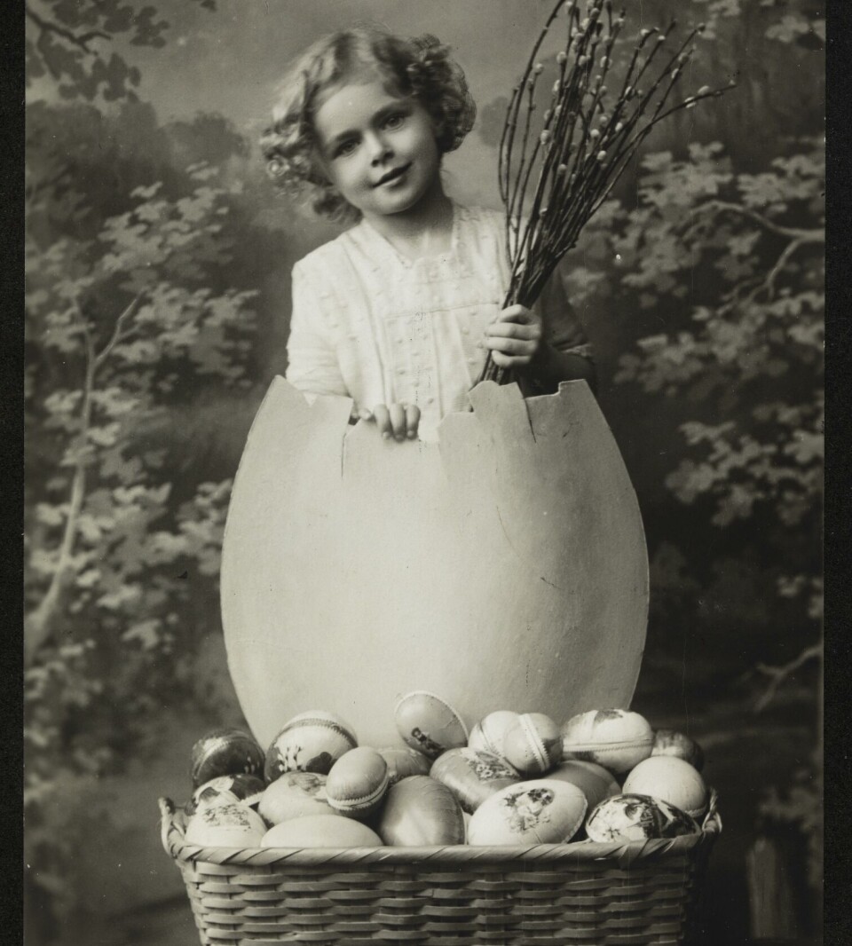 Dette fiffige påskekortet er fra rundt 1916. Et postkort i svart/hvitt som viser en liten jente som kommer ut av et stort egg. Ved føttene hennes står en kurv full av påskeegg.