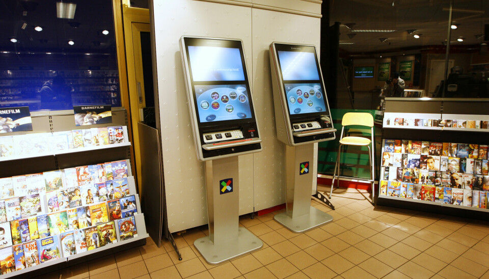 To spilleautomater i en norsk kiosk. Skjermene er blå med uklare, runde symboler. Automatene er merket med logoen til Norsk tipping. På siden av automatene er det hyller med blader.