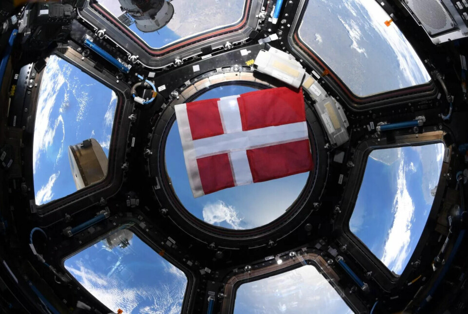 Det røde og hvite danske flagget ligger inntil et rundt vindu. Det er seks vinduer rundt dette som sammen danner en sirkel. Utsikten er blå med hvite skyer.
