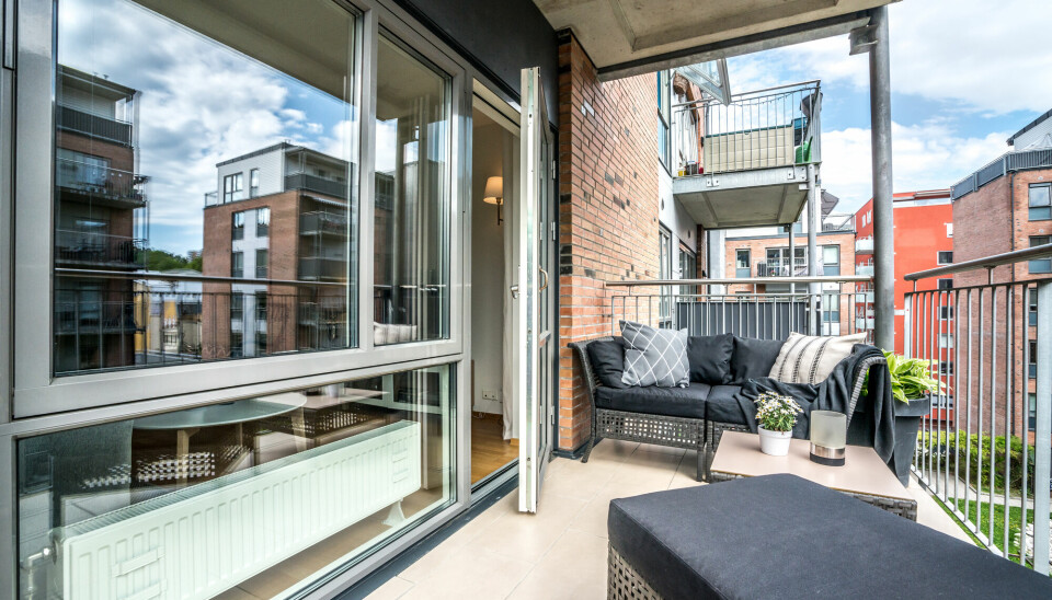 Balkong med rottingsofa og puff med svarte puter utenfor en leilighet med store vinduer i en nyere mursteinsbygård.