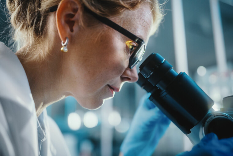 Nærbilde av kvinne på rundt 40 år med oppsatt, mørk blondt hår, perleøredobb og briller som ser inn i et mikroskop.