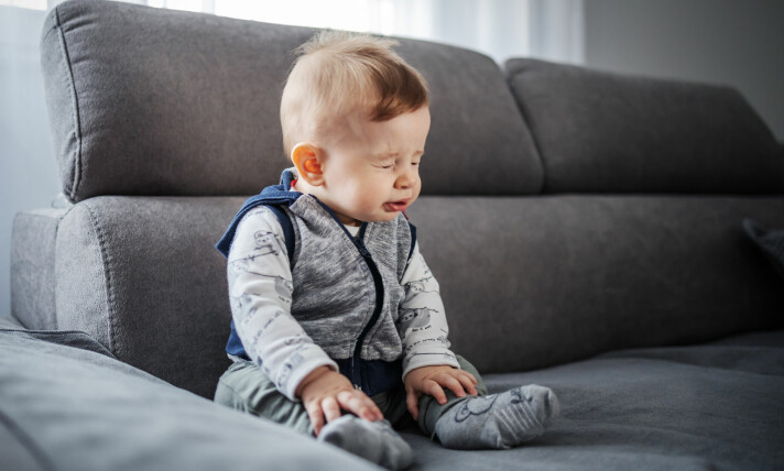 Liten babygutt med brunt hår sitter i en grå sofa og nyser.