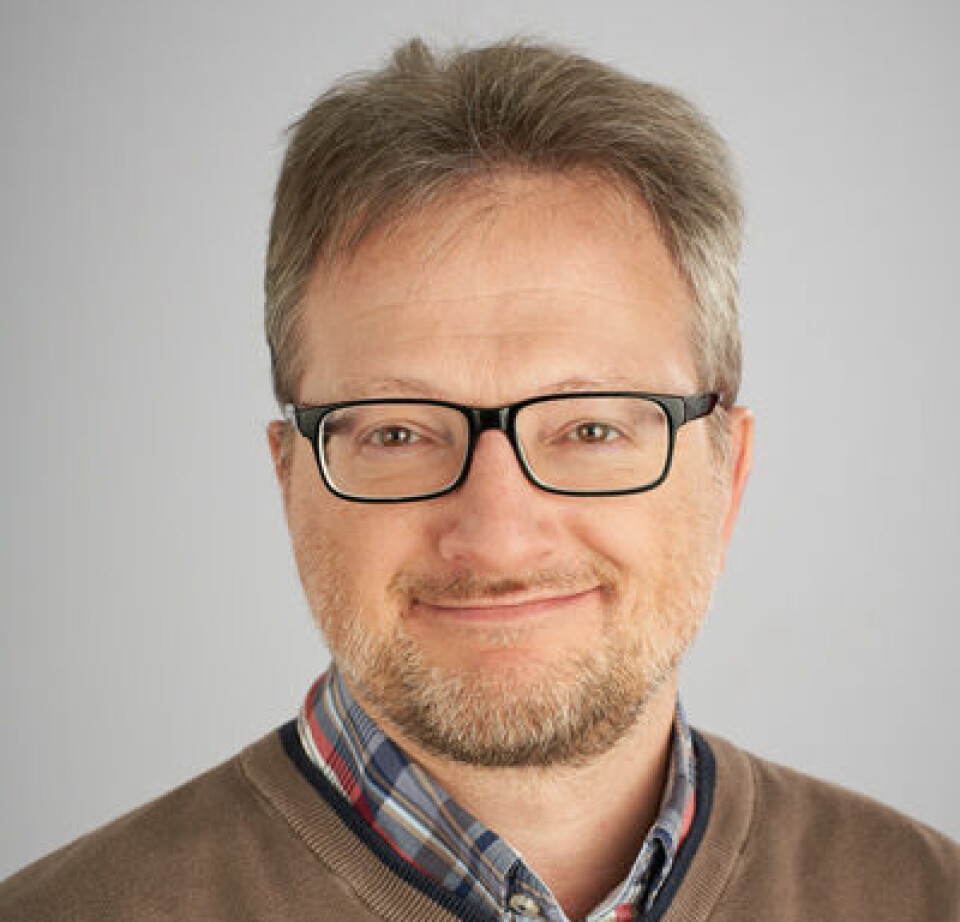 Portrettbilde av mann med briller, skjorte og brun genser som smiler til kamera.