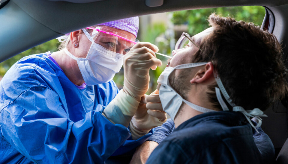 En mann sitter i bilen sin. En kvinnelig helsearbeider med maske, visir og hansker bøyer seg inn vinduet og stikker en bomullspinne opp i mannens nese.
