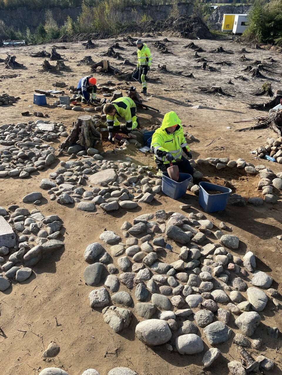 Oversiktsbilde av arkeologer som jobber på bakken. Steinsirkler på bakken markerer graver arkeologene undersøker.