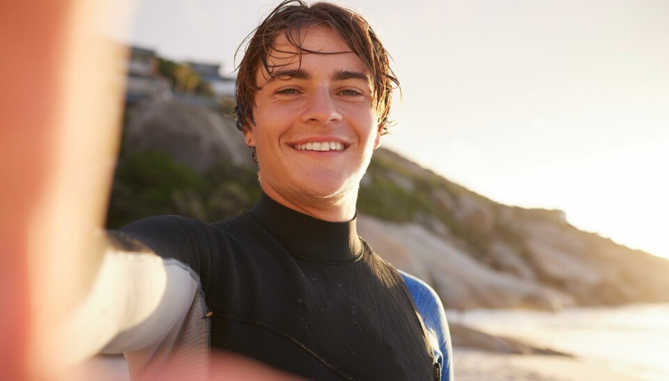 Tenåringsgutt med vått hår og våtdrakt tar selfie på en strand.