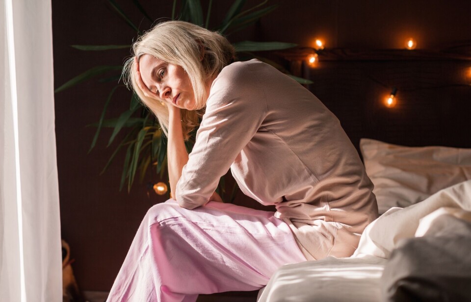 Når østrogennivået faller, kan det muligens utløse symptomer eller forverre allerede depressive symptomer. Middelaldrende kvinne med lyst, rett hår til rett over skuldrene sitter alvorlig på sengekanten.