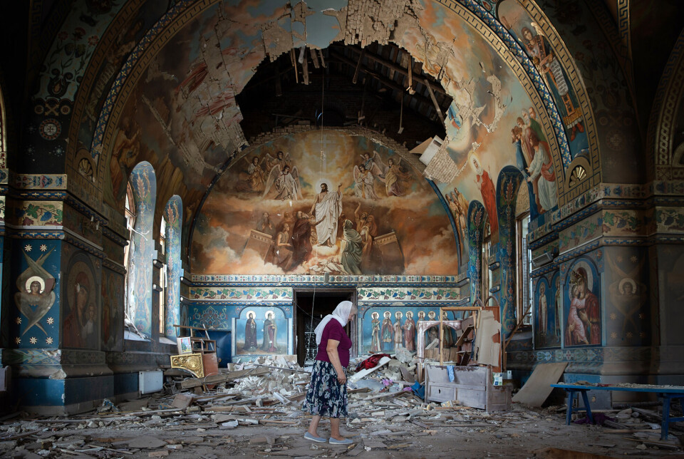 Lyudmilla Petrovna i kirken der hun selger lys og religiøse ikoner. Kirken i Novoekonomiche, ved Toretsk i Ukraina, ble skadet i et russisk angrep. Bildet er fra august 2024. Kirke med malerier og utsmykning hovedsakelig i brunt og turkis. I det buede taket er det et stort hull, rett over hodet på den eldre kvinnen med vinrød T-skjorte, mønstrete skjørt og hvitt skaut.