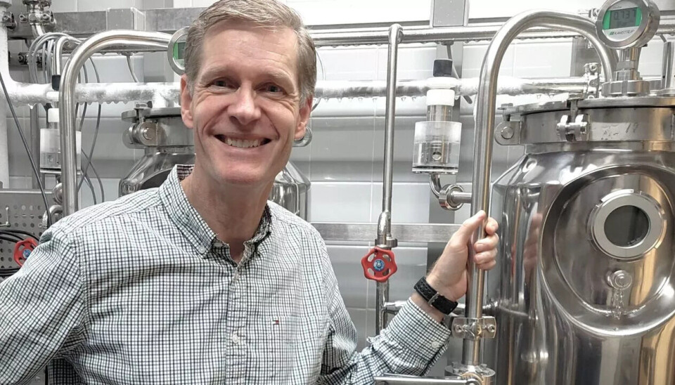 Bryggmester Erik Lund fra Carlsberg Laboratorium viser fram utstyret de bruker til å teste nye gjærtyper som lager øl uten alkohol. Smilende mann med lys blondt hår med gråstenk og smårutete skjorte i hvitt, grått og svart. Han holder i en blank metallstang festet til en metalltank.