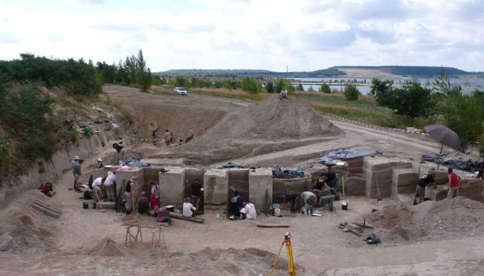 Ved det arkeologiske området Neumark‑Nord 2 i Tyskland har forskere funnet bevis for at neandertalerne var smartere enn vi trodde. En gruppe arkeologer graver ut et stort åpent område med dype grøfter og jordstolper, omgitt av trær, åser og en vannflate under en delvis overskyet himmel. Utstyr og redskaper ligger spredt rundt i området.