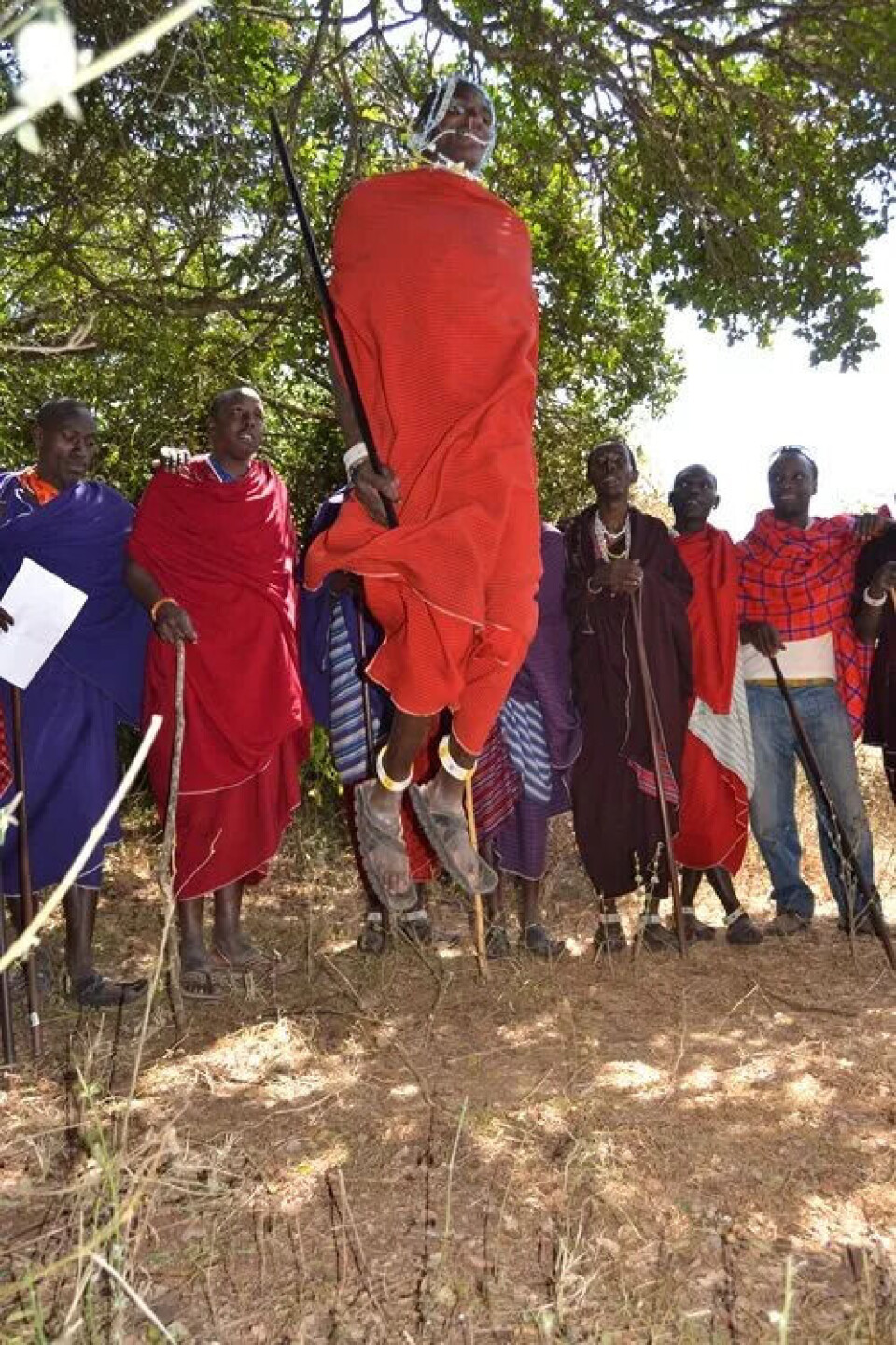 Ifølge kultursosiolog Niels Larsen bruker masai-mennene hoppesessionen som et slags flørtende datingritual. Mann i rød drakt og med en stav i hånda hopper. Han har sandaler på føttene. Flere menn står bak og ser på.