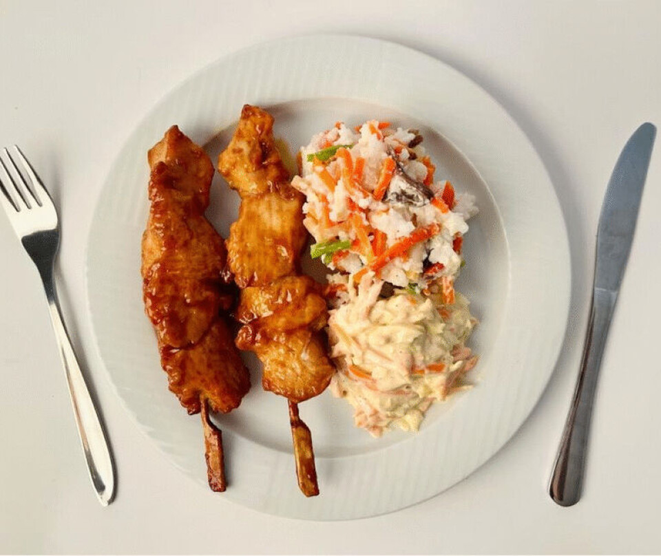 Ultraprosessert middag: Teriyaki-kyllingspyd, hvit ris med grønnsaker og coleslaw. Stilisert bilde på hvit bakgrunn av en hvit tallerken med middag. Til venstre på tallerkenen er to kyllingspyd med saus og til høyre en stor klump ris med strimler av gulrot og en grønn grønnsak. Gaffel til venstre og kniv til høyre for tallerkenen.