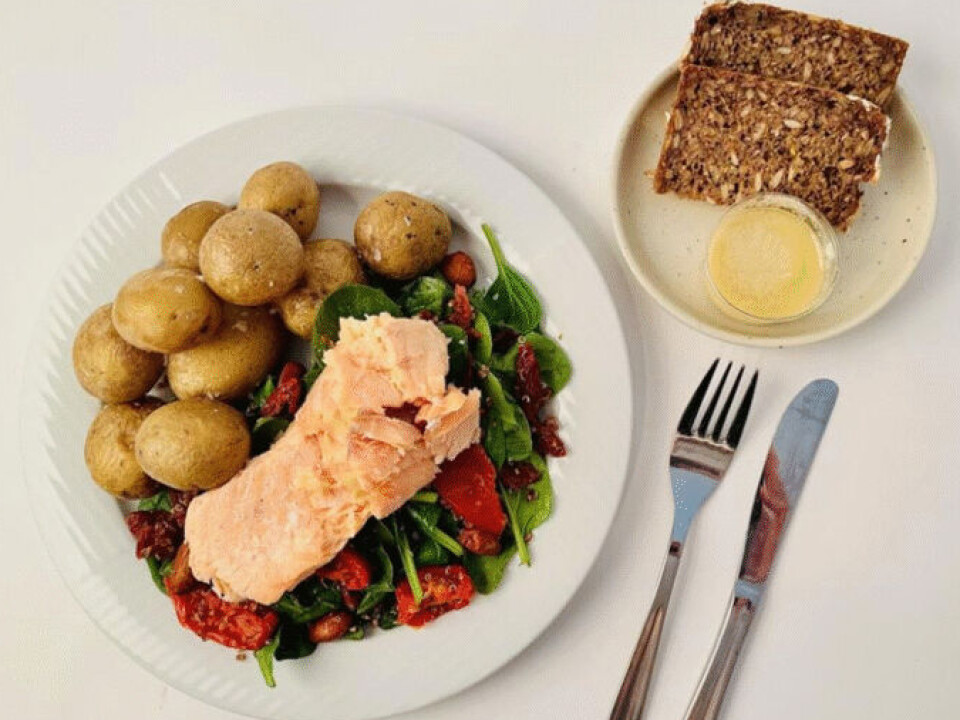 Minimalt prosessert middag: Quinoa- og spinat-salat, poteter, laks, rugbrød og appelsindressing. Stilisert bilde med hvit bakgrunn. Til venstre en hvit tallerken med ti poteter og et stykke laksefilet som ligger oppå spinat, quinoa og tomater. Til høyre en liten, hvit asjett med to skiver rugbrød og en liten glasskål med gulhvit saus. Kniv og gaffel ved siden av.