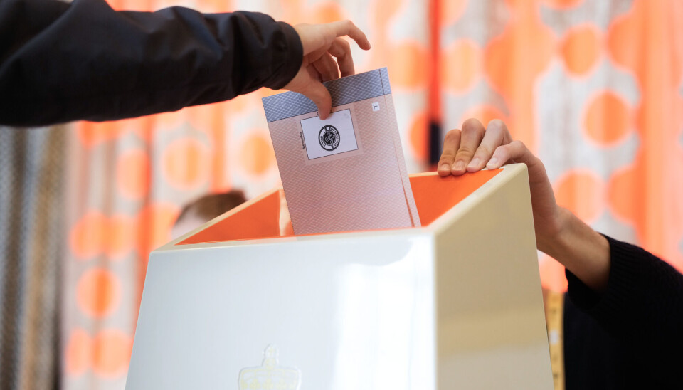 Kvinner og menn er ofte opptatt av litt ulike ting i politikken, og derfor stemmer de ofte på litt ulike partier. Hånd som putter stemmeseddel nedi en hvit stemmeurne som er oransje inni. En valgfunksjonær holder hånden på den andre siden av urnen. Oransje gardiner med hvitt mønster i bakgrunnen.