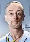 Middelaldrende mann med gråbrunt, tynt hår, skjegg og bart. Han har firkantede briller med tynn metallinnfatning og mørkebrune øyne. Ser inn i kameraet med oppsperrede øyne og et alvorlig ansiktsuttrykk. Har på seg lyseblå legeskjorte og et stetoskop rundt halsen.