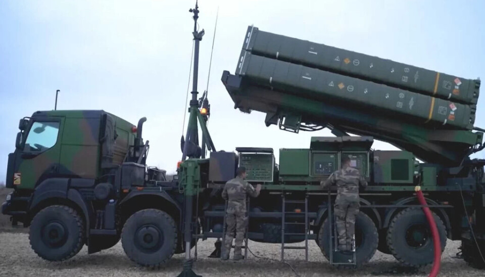 Danmarks nye missilsystem, det italiensk-fransk-produserte SAMP/T, kan også skyte ned droner. Det er et mobilt bakke-til-luft luftforsvarssystem. Her er en utgave fra det amerikanske forsvaret. To soldater i kamuflasjeuniformer arbeider med et militært missilsystem montert på en stor lastebil, med missilbeholdere hevet og utstyrspaneler åpne. Dette foregår utendørs på grus under en lett overskyet himmel.