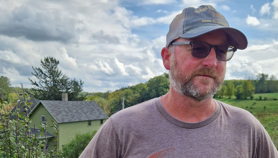 Nels Solum er syvende generasjon etter Niels og Margit Solum. Han driver gården i dag. Våningshuset i bakgrunnen er det andre huset familien bygde. Portrett av Nels Solum med våningshus i bakgrunnen.