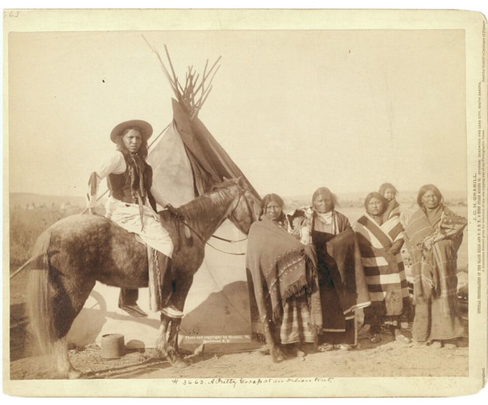 Folk fra Lakota-stammen i Pine Ridge-reservatet i 1891. Grådige agenter fra de føderale myndighetene tok selv penger og forsyninger som skulle gått til urfolket. Gruppebilde av mann og fire kvinner foran telt.