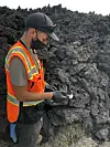 En mann i oransje refleksvest står med en liten hammer i hånden, foran en haug av mørk, knudret lavastein.