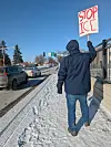 Person i vinterklær holder et «STOP ICE»-skilt ved en snødekt veikant med trafikk.