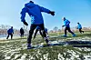 Barn trener fotball på snødekt kunstgressbane en kald vinterdag. De er iført klarblå, tynne treningsjakker og svarte treningsbukser.