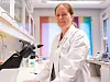 Professor og vaksineforsker Gunnveig Grødeland i hvit frakk står ved et Nikon-mikroskop i et lyst laboratorium.