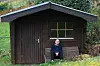 Mann sitter på benk foran mørkebrunt skur i grønne omgivelser