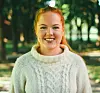 Ung kvinne med lys genser smiler utendørs i en allé med trær.