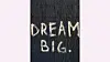 En hvit graffiti med teksten DREAM BIG er malt på en mørk, strukturert vegg.