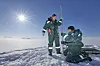 To forskere i vinterdresser måler snødybde på en solrik isbre.