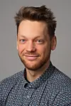 Smilende mann i mønstrete skjorte foran grå bakgrunn.