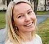 Smilende kvinne i lys genser står ute med bygninger og gress i bakgrunnen.
