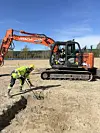 Et jorde med en gravemaskin og en arkeolog som holder en metalldetektor.