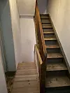 Smal innendørs trapp i tre med rekkverk i et trangt rom med lyse vegger.